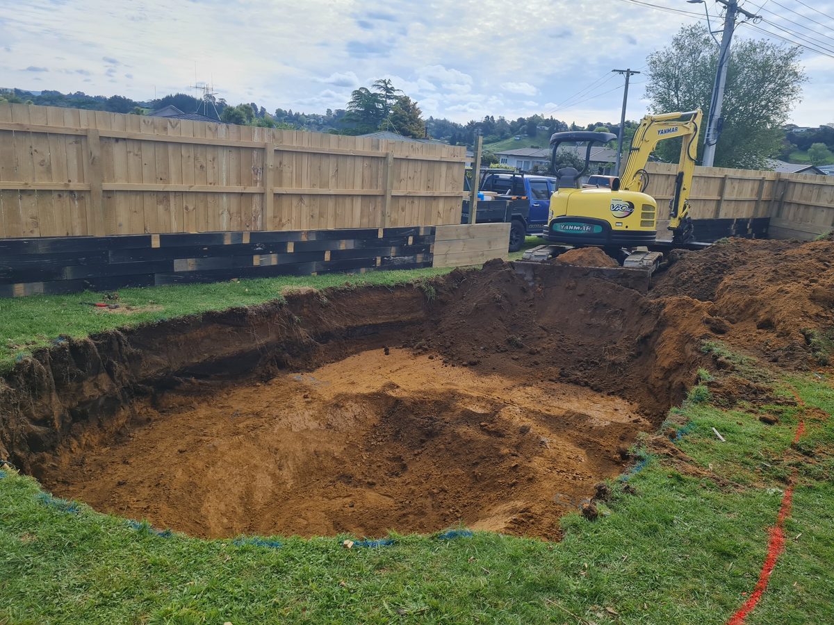 A digger beside a freshly dug pool excavation in a fenced back yard
