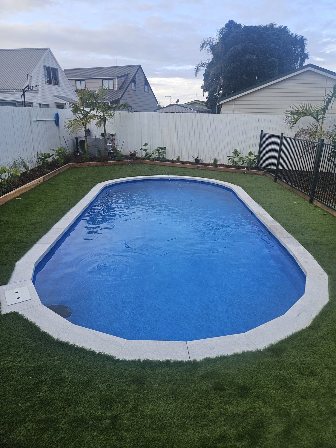 An oval pool with a white coping in artificial grass behind a glass fence