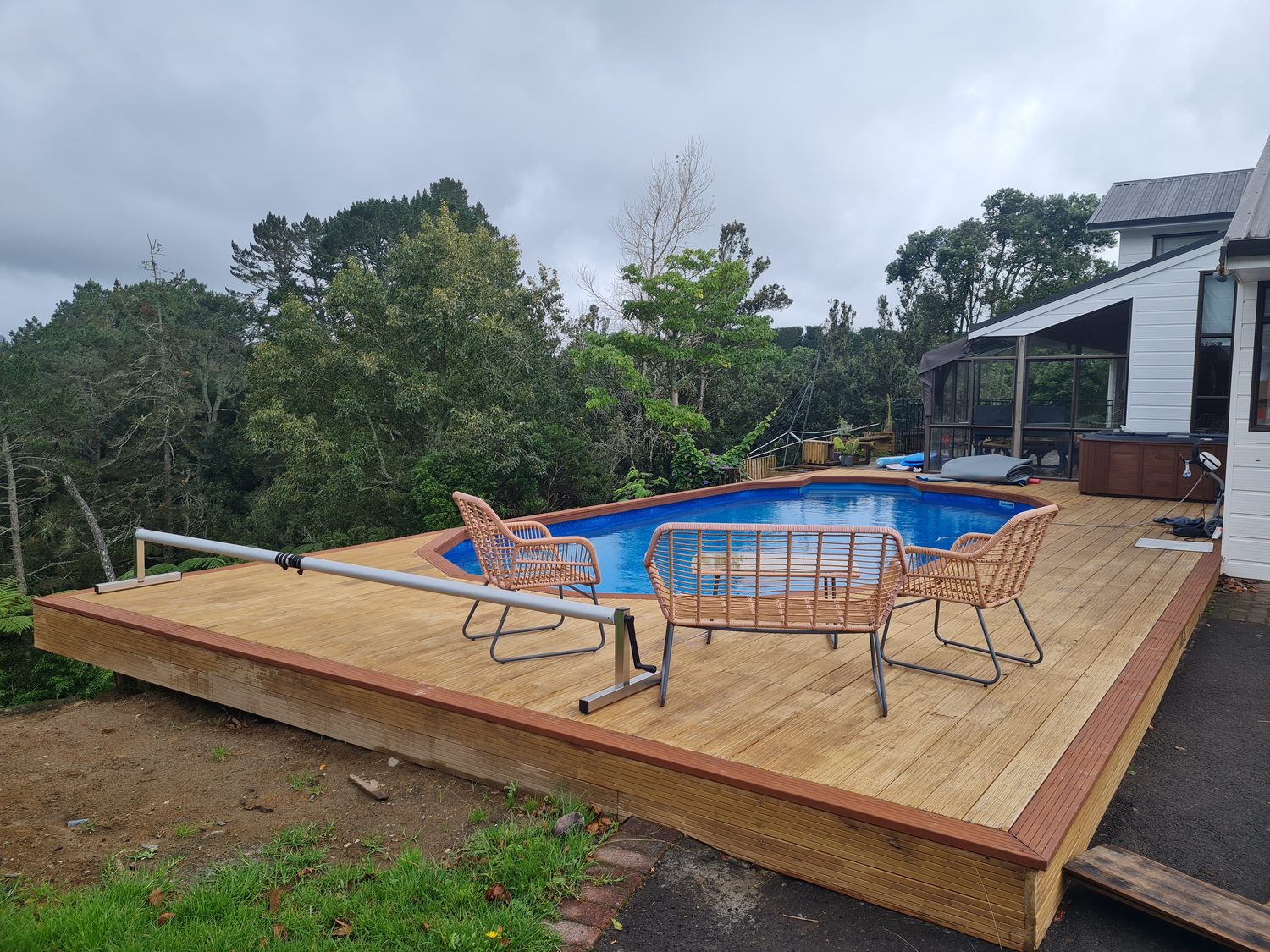 A pool in a raised timber deck with chairs and a cover roller
