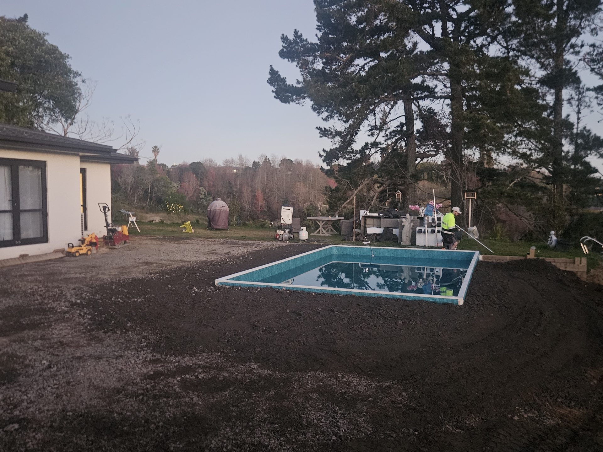 A newly built rectangular pool on bare ground beside a house at dusk