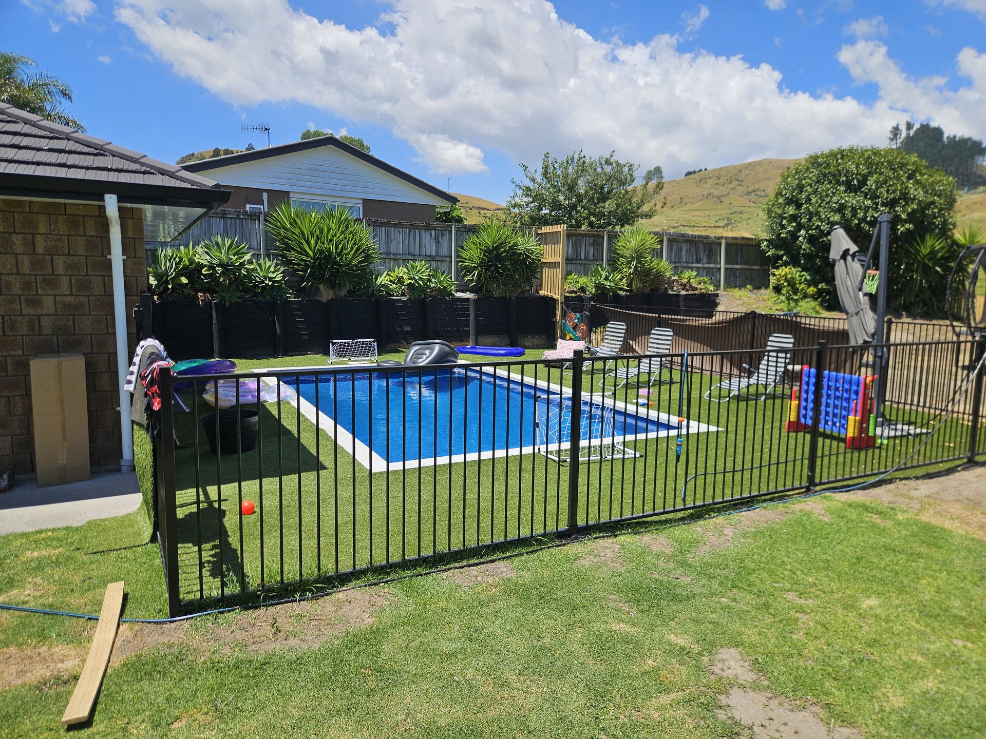 A rectangular pool on a fenced lawn behind a house, with pool toys on the grass